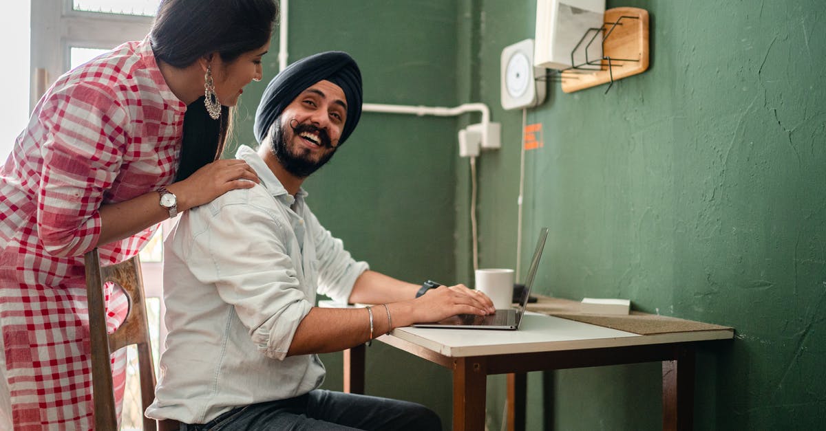 Dual National Travel back home using expired passport as Visa - Side view of ethnic male in casual clothes and hat using netbook at table while ethnic woman in casual clothes bending over and touching back of man