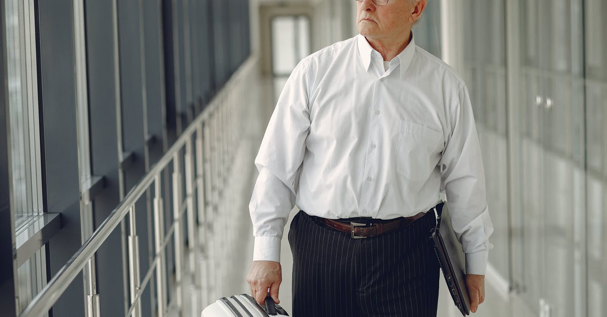 Electronic Devices and airport security checks [duplicate] - Serious senior male in formal clothes walking along airport corridor with baggage and laptop while looking away through window