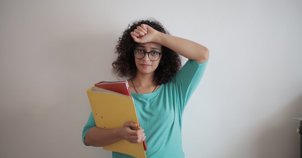 Emirates - Family bonus account with multiple frequent flyers - Unhappy overworked female employee in glasses and casual clothes with folders of documents looking at camera with sadness and touching head while standing against white wall in contemporary office