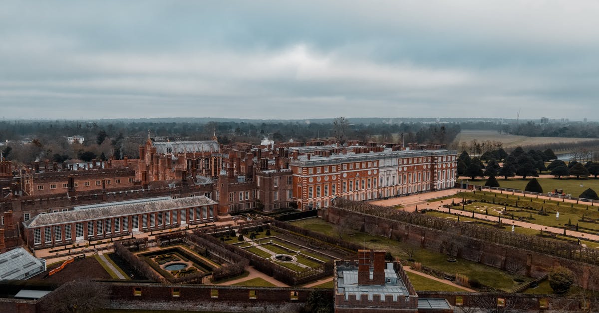 Extending 7 day London Oyster card for extra zones - Drone view of Hampton Court Palace located in England in London with green grassy lawns with plants near fence and trees near pathways with people under cloudy gray sky