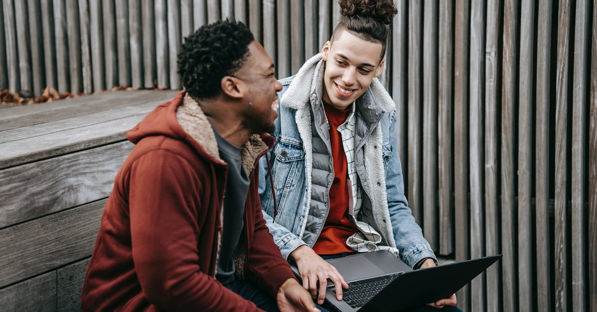 Extraordinary circumstances, missed connections and overnight stays - Cheerful multiracial students talking and smiling while surfing internet on netbook on wooden stairs near fence in daytime