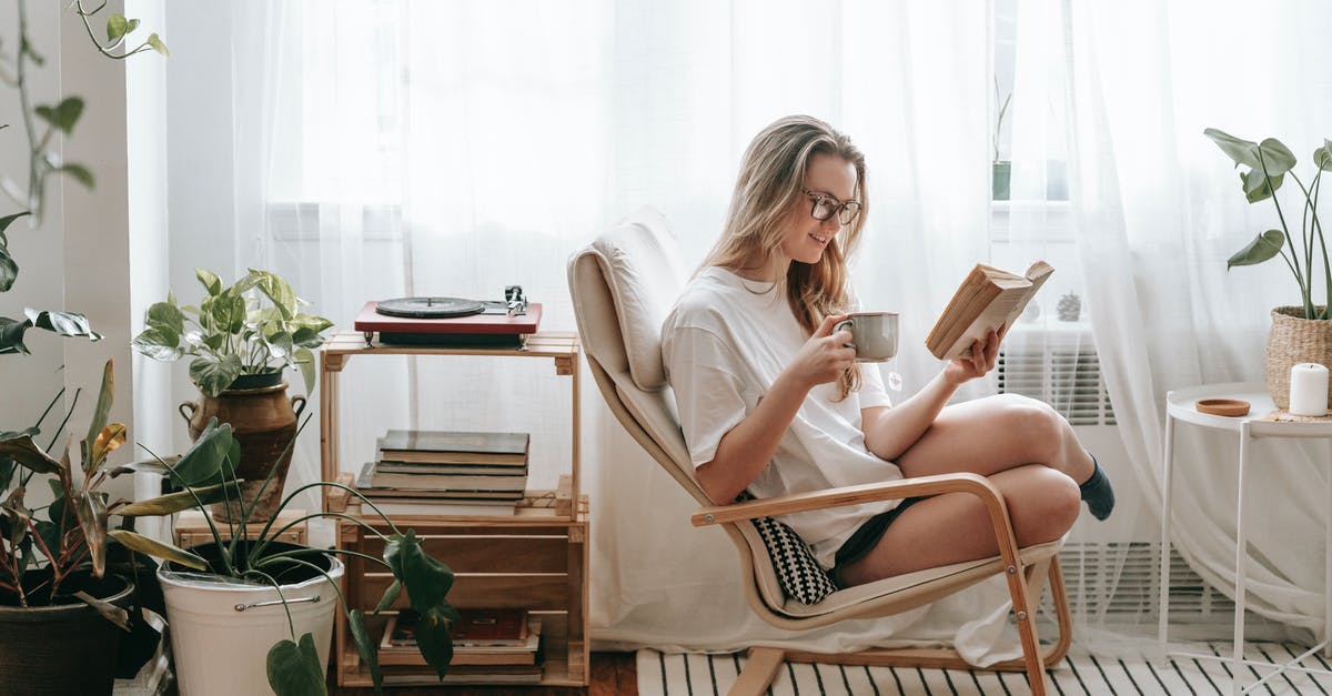 Free Amtrak Lounge Access with No Ticket - Cheerful young female in eyeglasses with cup of beverage reading textbook in armchair between potted plants in house room