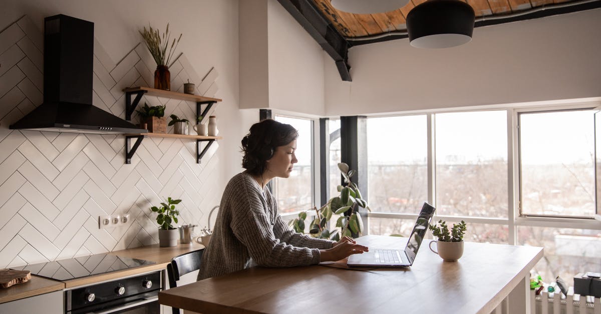 French student in Canada: going back home from NYC - Side view of young female in casual clothes typing message on laptop while sitting at wooden table in kitchen