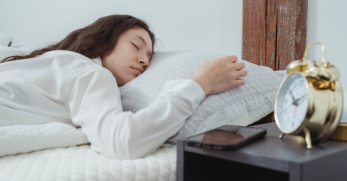 Getting to Reagan Airport (DCA) early in the morning - Young woman with dark long wavy hair sleeping peacefully on belly on comfortable bed under white blanket near bedside table with alarm clock and smartphone