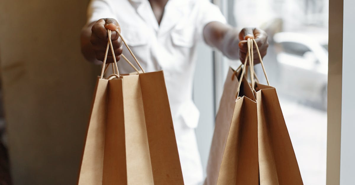 Give Delta voucher to another person - Crop anonymous African American person in white jacket passing purchases in paper shopping bags while standing near window in daylight