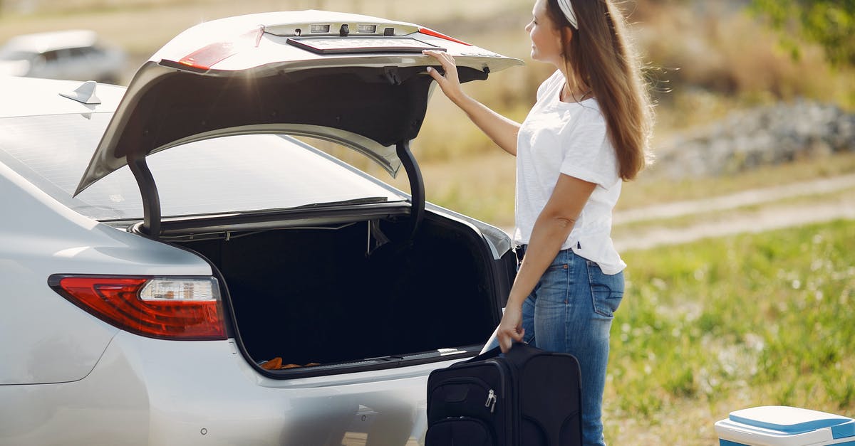 Hard-case suitcase, that doesn't open at the side - Side view of cheerful female traveler in headband and casual clothes putting luggage in open trunk of modern car while spending summer weekend in countryside