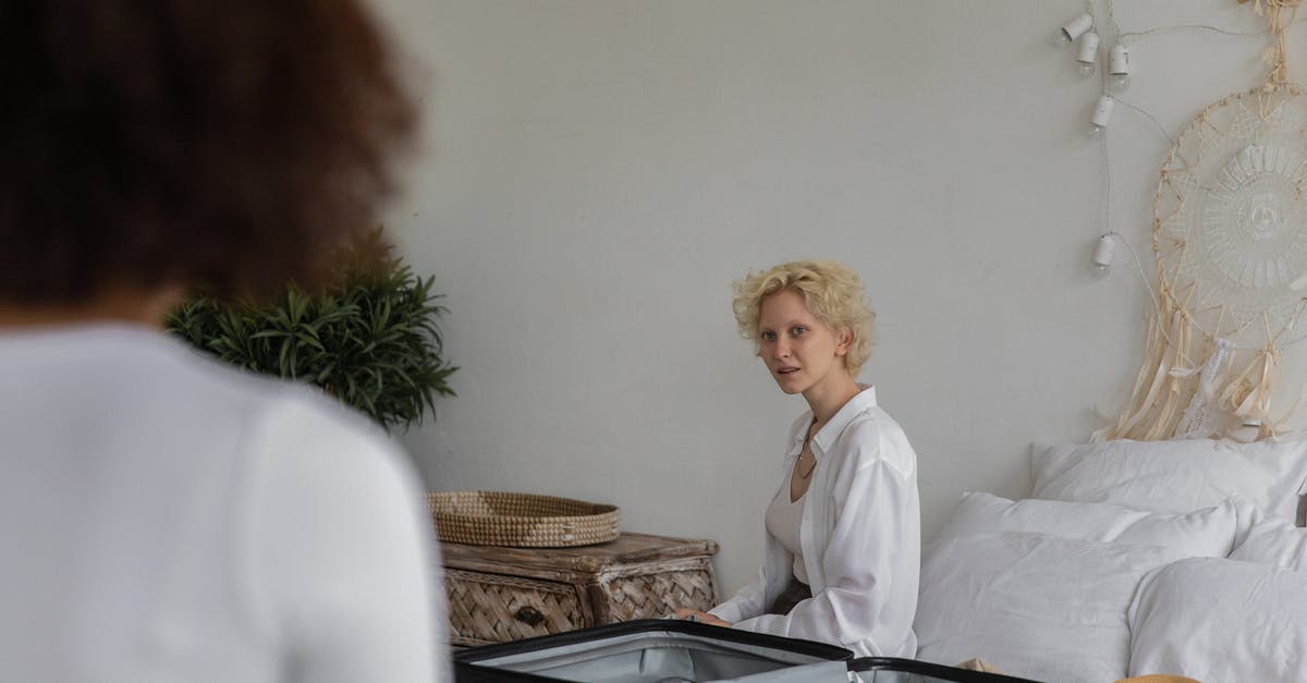 Helping friend with luggage at London Euston - Content lady with blond hair preparing to leave while sitting on bed near white wall in bedroom with crop female friend with dark hair