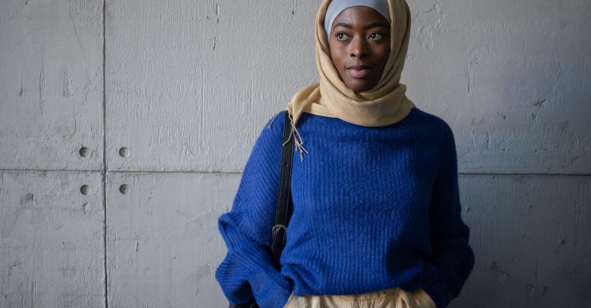 Hitchhiking through Native American reservations - Content Muslim female student in traditional headscarf wearing blue sweater looking away while standing near wall with backpack in corridor of university