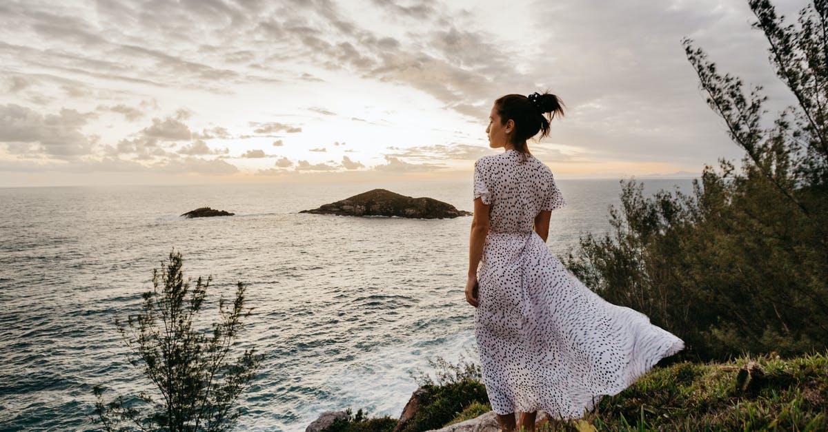 Holidays in Brazil without Portuguese - Back view of calm young female traveler in elegant summer dress admiring seascape from grassy cliff against cloudy sunset sky in Arraial do Cabo