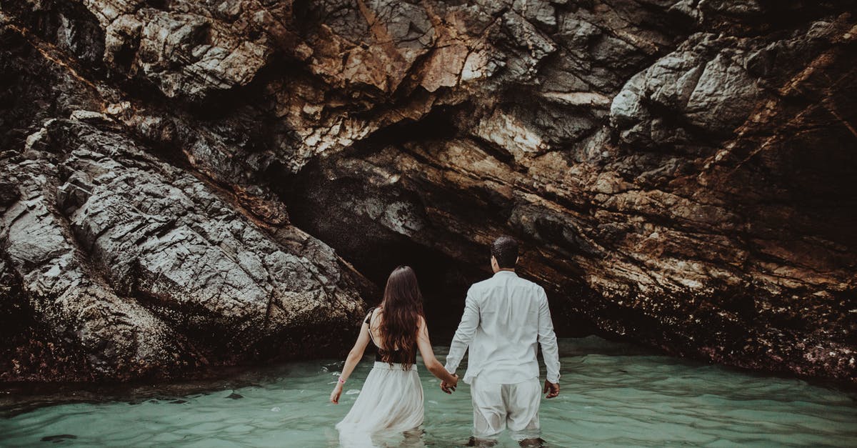 Honeymoon destination options [closed] - Back view of unrecognizable romantic couple in summer clothes holding hands and walking in sea water near massive rocky cave