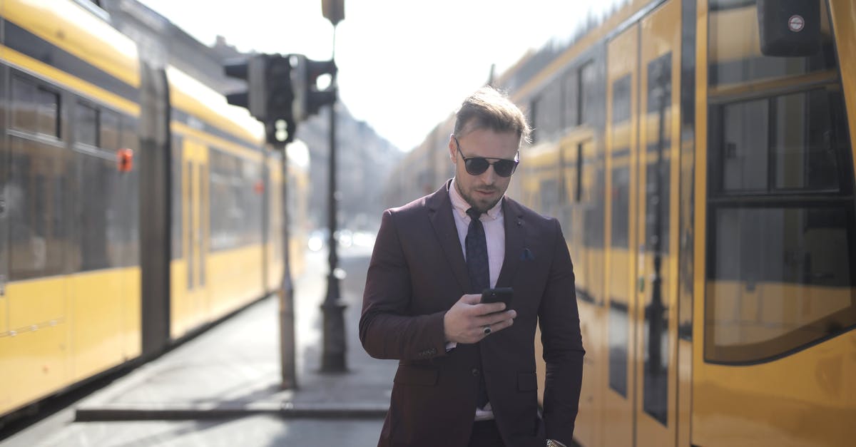 How can I work out the price of a UK bus fare online? - Brutal male entrepreneur in elegant suit and sunglasses standing with hand in pocket on street between trams and messaging on cellphone