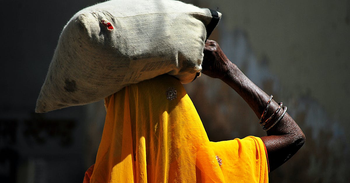 How do airlines compute weight allowances for carry-on bags? - Back view of unrecognizable African female in traditional yellow headscarf carrying heavy sack on head while walking in village on sunny day