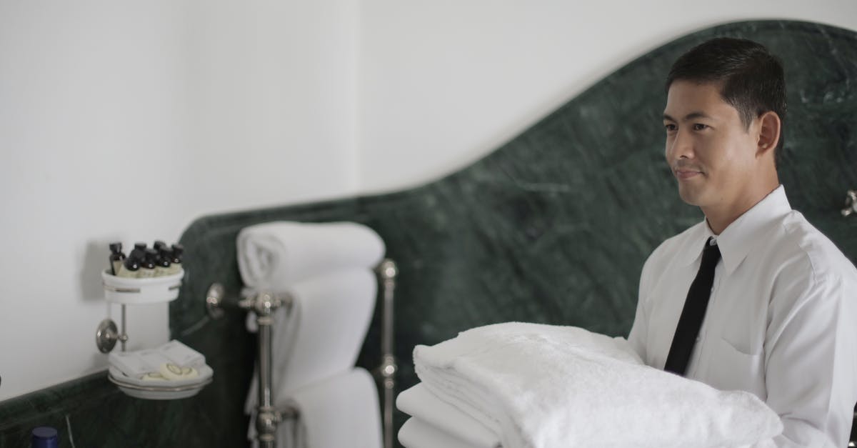 How do hotel housekeepers know if a room is in use? - Side view of smiling ethnic male hotel employee holding stack of clean white towels while cleaning bathroom