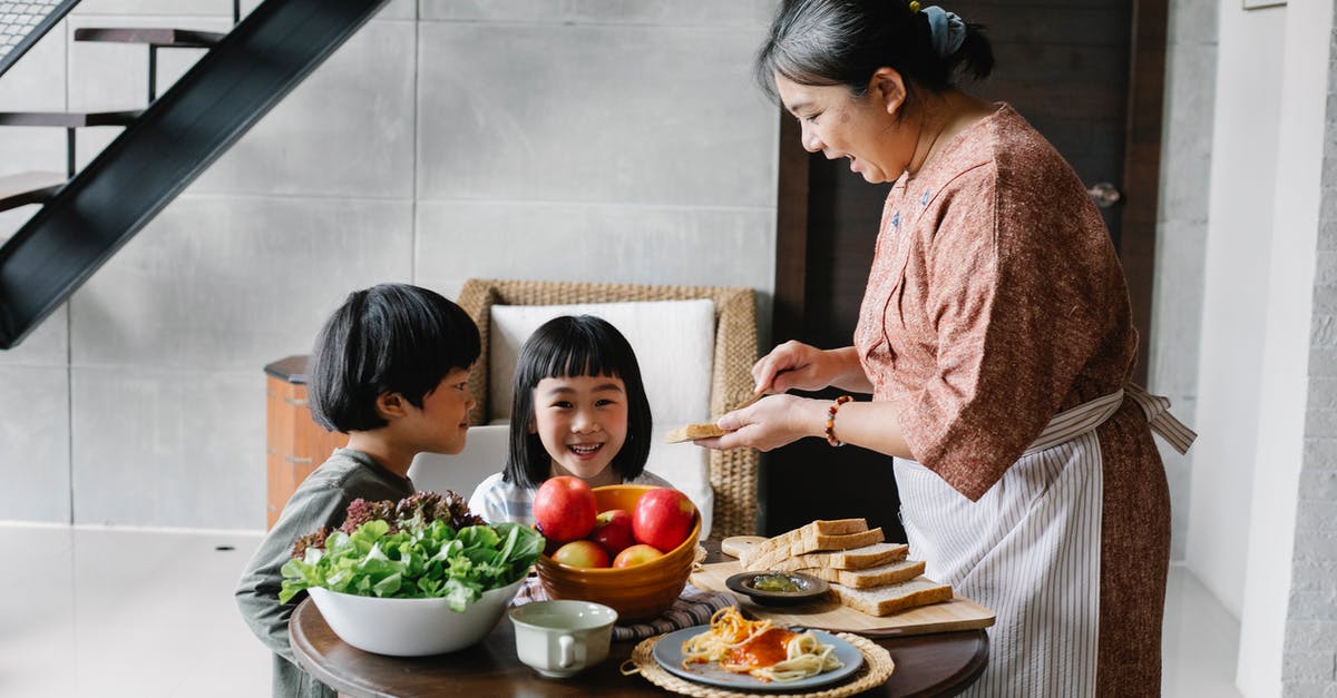 How do I make sure I'm not awakened during in-flight food service? - Side view of positive senior Asian female in apron spreading butter on bread while preparing breakfast for funny little grandchildren sitting at round table in cozy kitchen