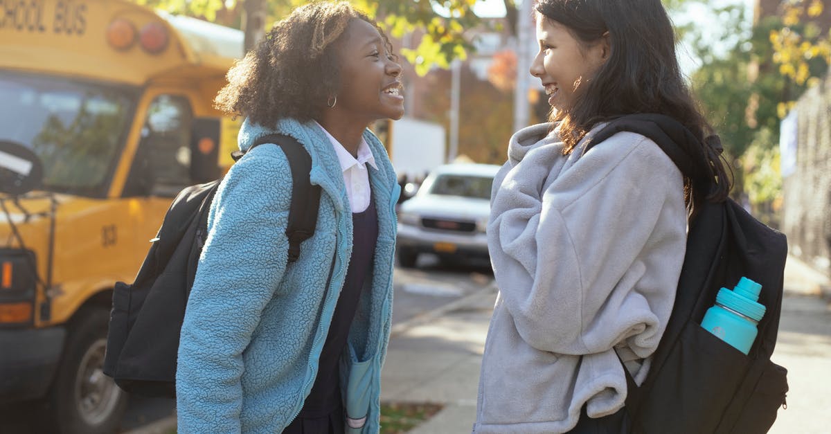 How do US intercity bus companies relate to each other? - Side view happy multiracial schoolgirls in warm jackets with backpacks standing on street near school bus and talking while looking at each other with smile