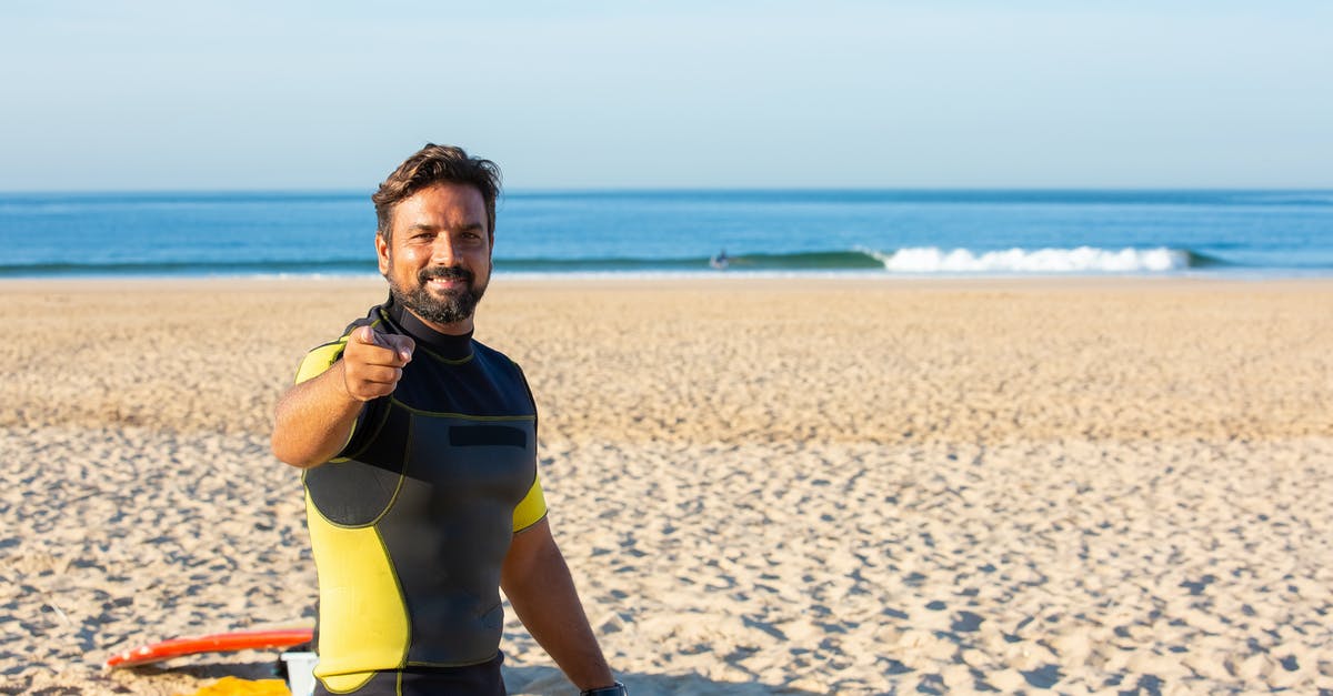 How do you choose a restaurant when travelling? - Happy male in diving suit looking and pointing at camera while standing on beach in bright sunny day