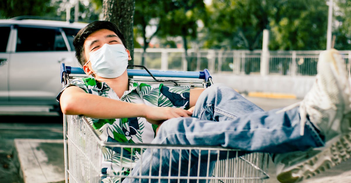 How do you keep a sleep mask on while asleep? - Young Asian male in casual summer wear and face mask lying with eyes closed in shopping trolley on sunny street