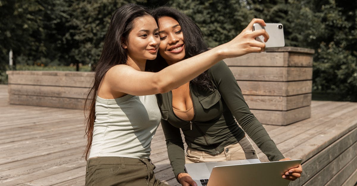 How long time does it take to renew a Schengen visa? - Positive young multiethnic ladies with long dark hairs in casual outfits smiling and taking selfie on mobile phone while chilling in park with laptop