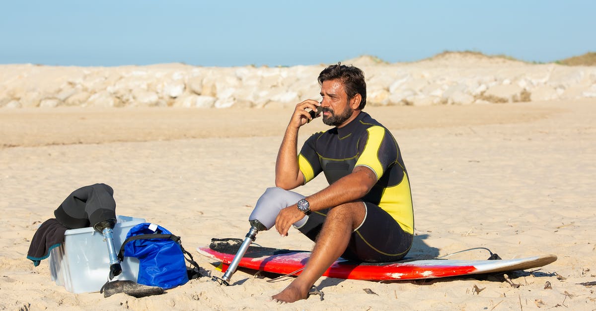 How to activate mobile phone in Chile? - Full length tanned bearded male surfer with leg prosthesis sitting on sandy beach on surfboard and having conversation on mobile phone
