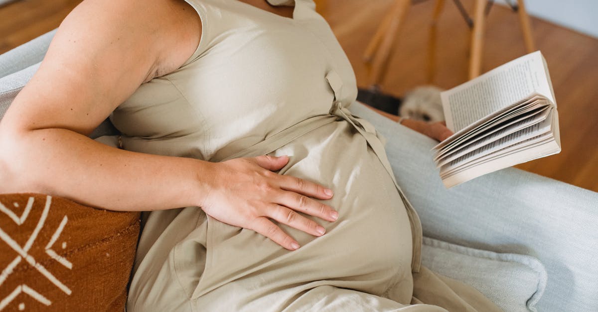 How to book a ticket for an unborn infant? - Faceless pregnant female in beige dress reading book on comfortable sofa and touching tummy