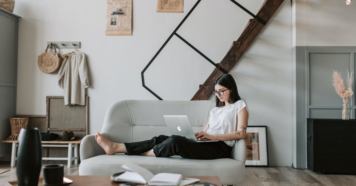 How to book a train ticket from Tblisi to Baku (online) - Positive female with tattooed arms wearing casual clothes and eyeglasses reading netbook while sitting barefoot on comfortable sofa behind table with books and magazines in modern apartment with minimalist interior in eco style during free time