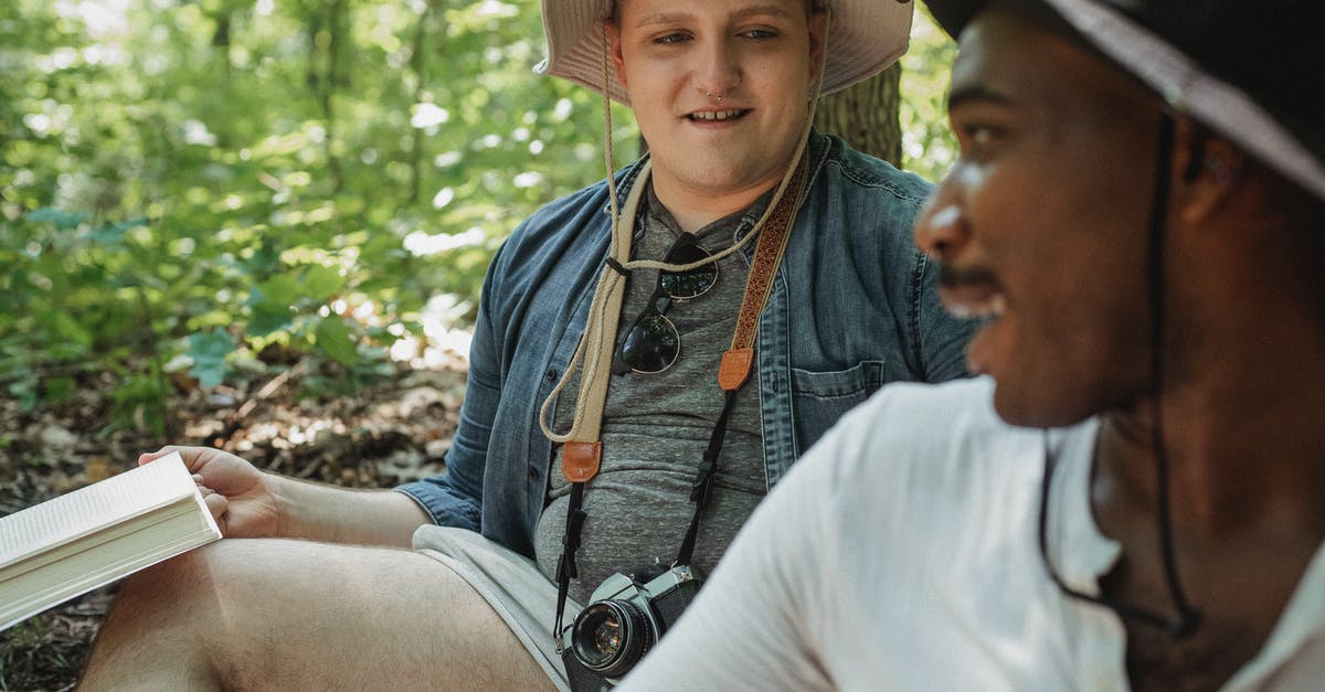 How to book flights when travelling together with slightly different itineraries? - Smiling bearded African American man in black hat looking over shoulder on man while sitting together on blurred background in woods