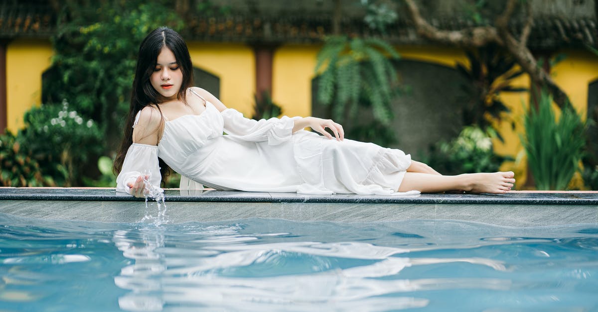 How to clean drug misdemeanor in US so I don't get stopped at border every time? - Full body of thoughtful barefoot Asian female in white dress looking down while lying on poolside near clear water near building and tree
