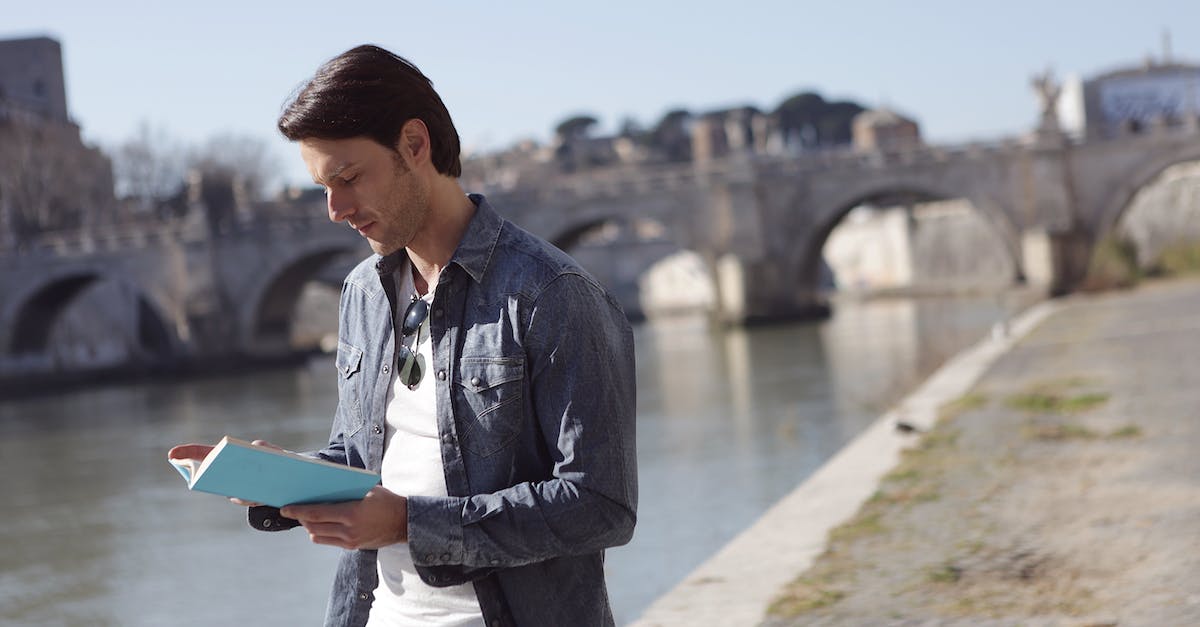 How to document travel expenses? - Young Man in Denim Jacket is Reading a Book Near River Side