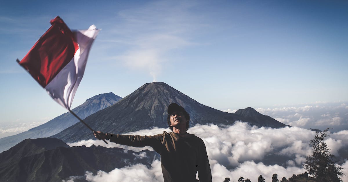 How to extend Indonesian visa on arrival (VOA) in Indonesia - Man Wearing Black Crew-neck Sweater Holding White and Red Flag Standing Near Mountain Under Blue and White Sky