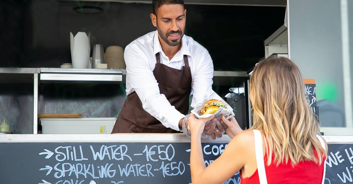 How to order food on Zakynthos - Positive ethnic cook in apron standing at counter in food truck and giving delicious hamburger to anonymous woman customer in daytime