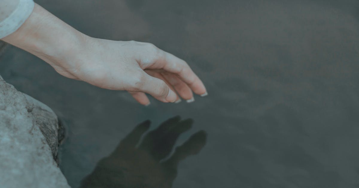 How to reach the clearest lake in the world? - Crop anonymous female on boulder on coast near pond touching peaceful water with hand in daylight