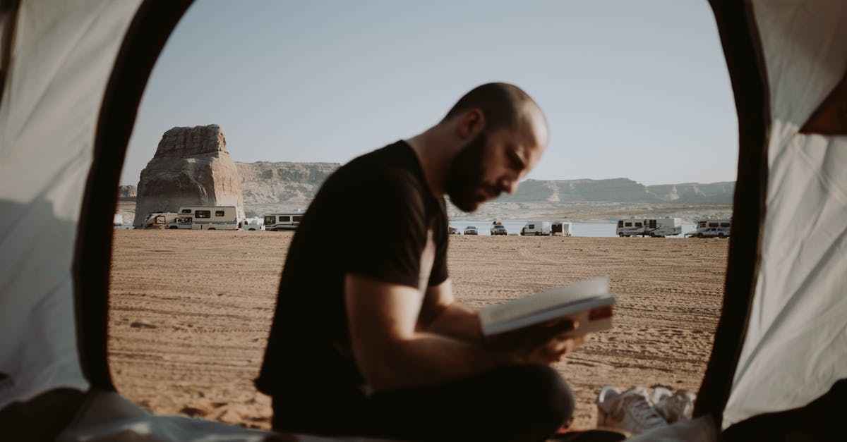 How to register while wild camping in the Balkans? - Side view of bearded male traveler sitting in tent with opened door and reading book in nature with parked cars in distance