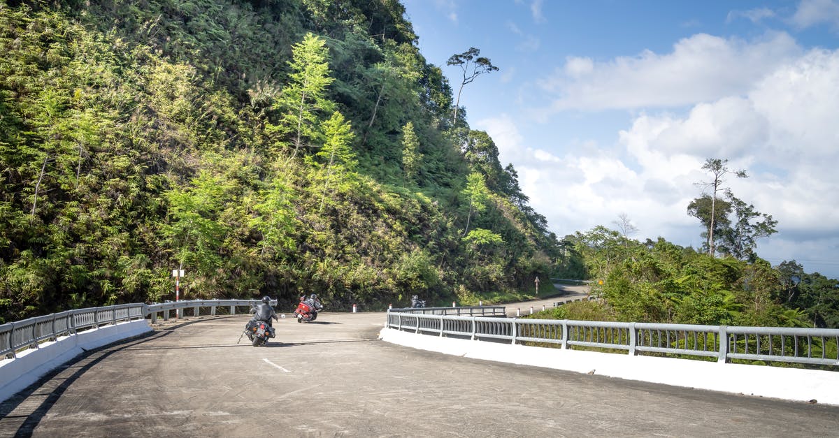 How to safely transport external hard drives during air travel? - Back view of unrecognizable motorcyclists driving bikes on asphalt roadway with fences against lush trees on mount under cloudy blue sky