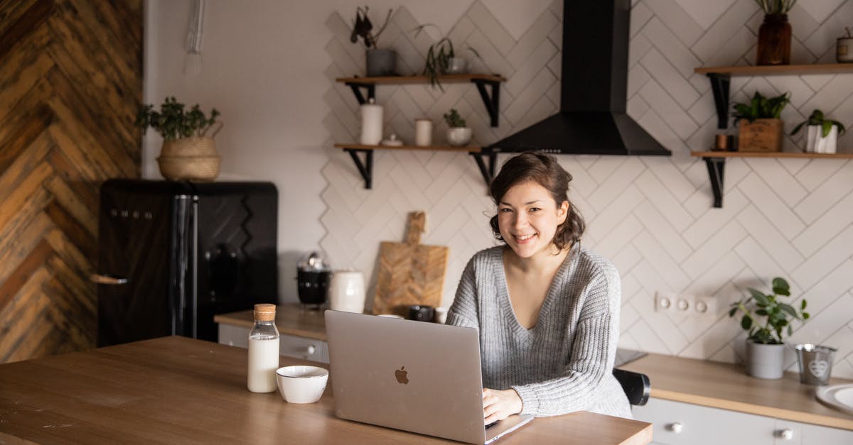 How to search for weekend flights? - Cheerful female in casual clothes sitting at table with laptop and bottle of milk while browsing internet on laptop during free time at home and smiling looking at camera