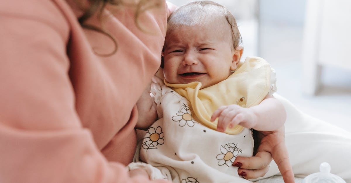 How to sterilize infant feeding bottles while traveling? - Crop anonymous caring mother with crying newborn baby in arms sitting on couch in light room with feeding bottle at home