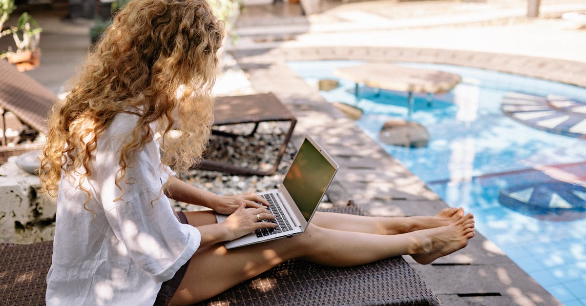 How useful are bitcoins when it comes to traveling? - From above side view of unrecognizable barefoot female traveler with curly hair typing on netbook while resting on sunbed near swimming pool on sunny day