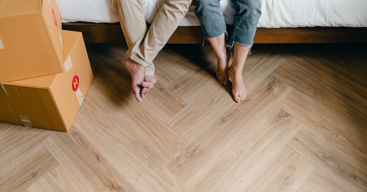 Husband and Wife TSA PRE-Check Changes - Unrecognizable barefoot man and woman sitting on bed near carton boxes while relaxing from unboxing belongings during relocation in new flat