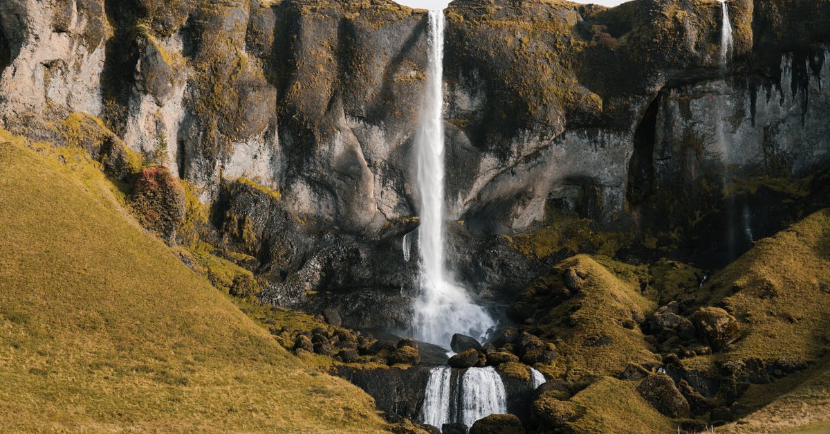 Iceland's Schengen Area situation - Picturesque distance scenery of small waterfall falling off tuffstone cliffs and green slopes with cattle grazing at foot of mountains during daytime