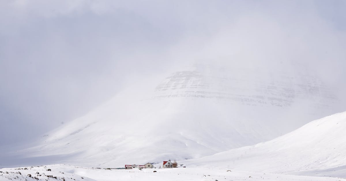 Iceland - Visiting fjords from Reykjavík in 1 day - Small settlement in remote cold valley at bottom of big snowy mountain with peak in hidden in haze in sunny winter day