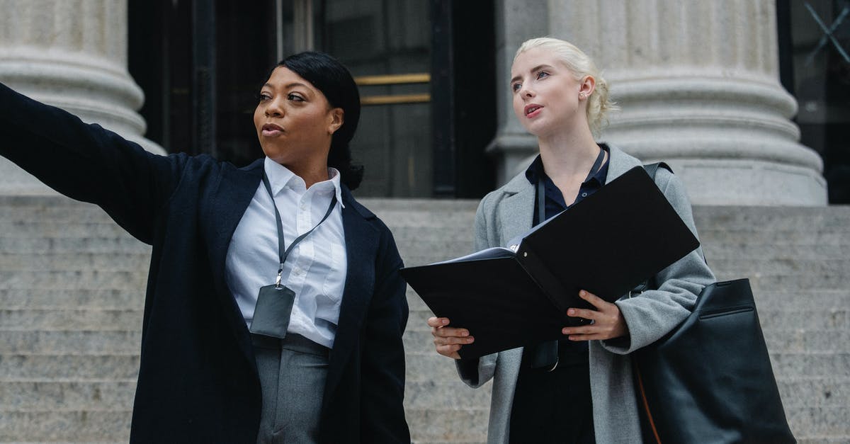 Identify the city from a Microsoft Teams meeting's background - Low angle of African American lady formal outfit and badge pointing away while standing on street with female colleague with documents folder