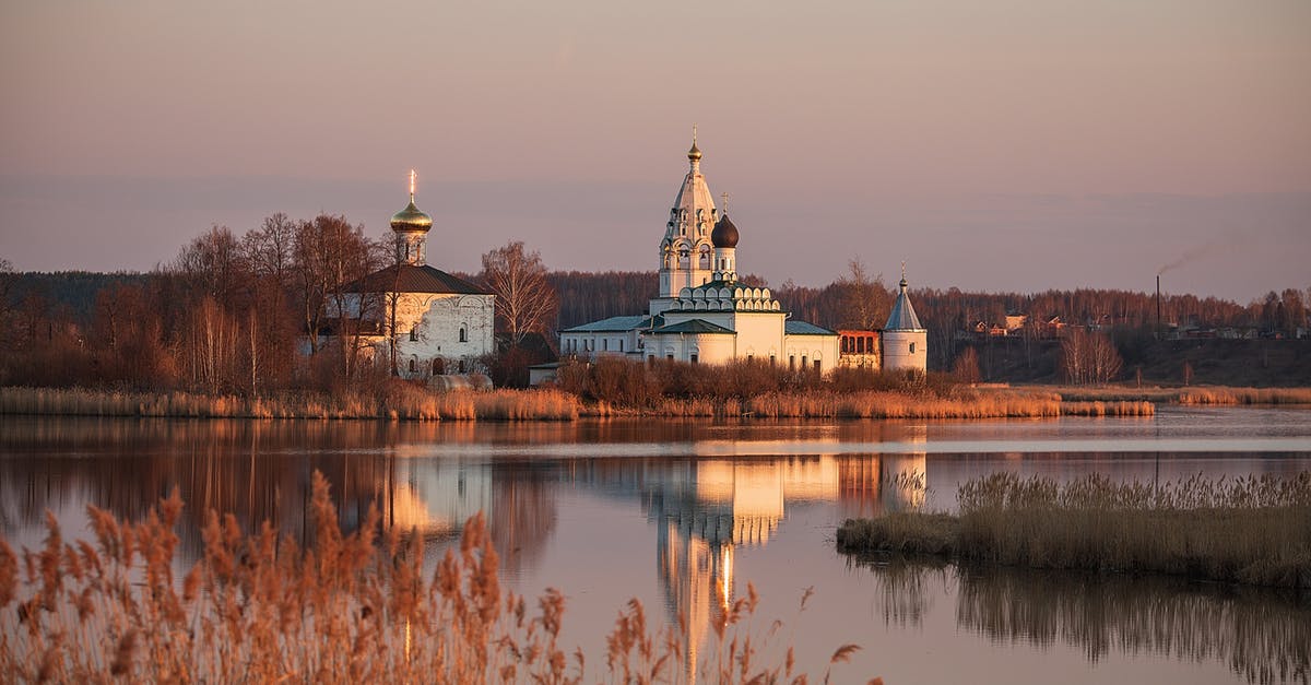 Identify waterside building / skyline - Scenery view of church with domes and withered trees located on shore of calm lake against cloudless sky in sunset