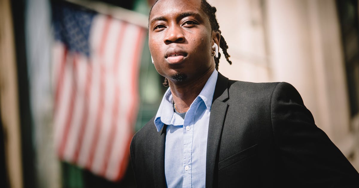 Indian citizen visiting Germany from US - Serious black businessman against blurred building with American flags