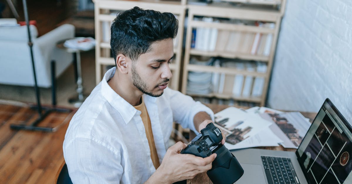 Indian e-visa photo rotated left (270 degrees) - Side view of Indian male photographer in casual clothes watching photos on photo camera while sitting at table and browsing laptop at home in daytime