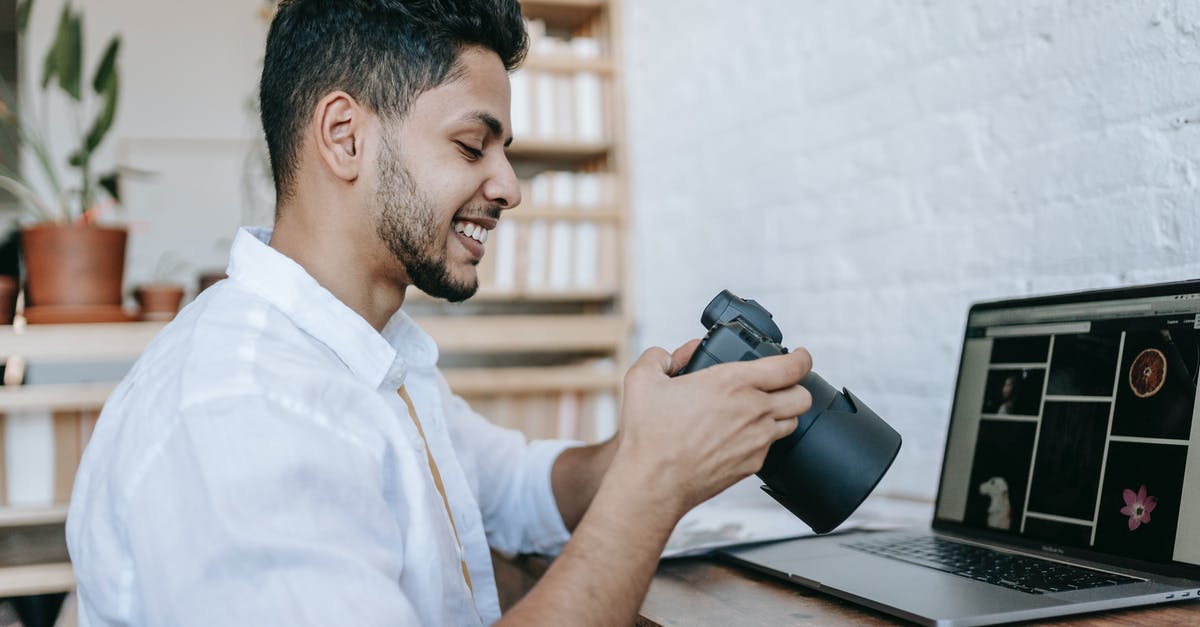 Indian e-visa photo rotated left (270 degrees) - Side view of young ethnic happy guy in white shirt with professional photo camera near wooden table with netbook and photos in bright apartment