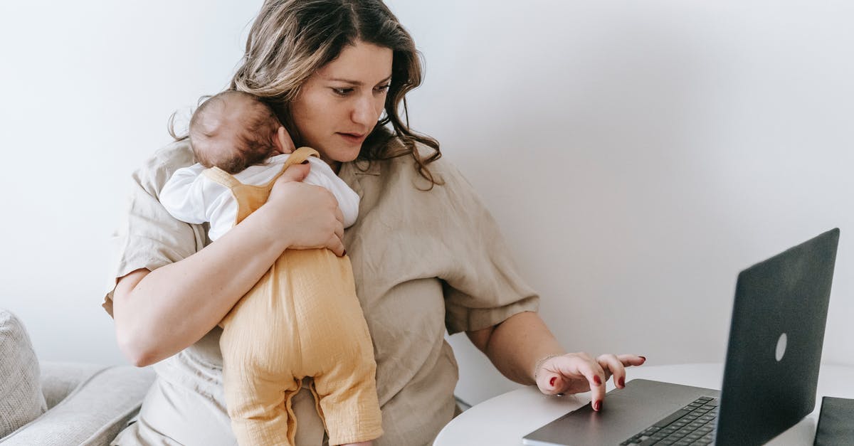 Ireland visit visa for work permit holder parent - Concentrated young female freelancer embracing newborn while sitting at table and working remotely on laptop at home