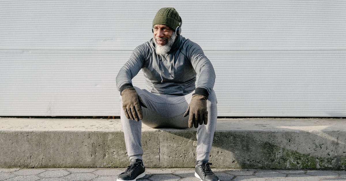 Is appearance important at the border? - Full body of positive African American male wearing trendy outfit listening to music with headphones while sitting on concrete border of road