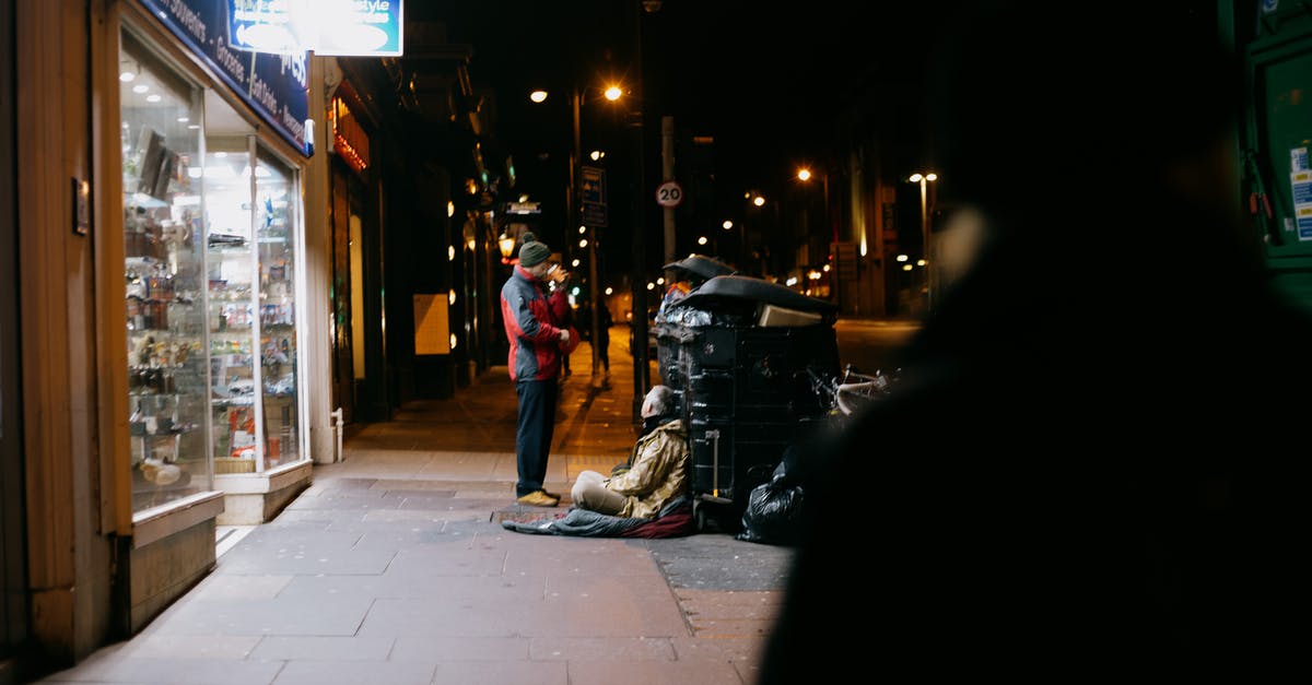 Is it bad to give money to beggars in India? - Anonymous man giving money to poor beggar on tiled sidewalk near shop showcases in town at dusk