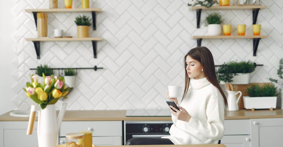 Is it inappropriate to record the call to prayer? - Side view of focused young woman in warm sweater browsing mobile phone in cozy kitchen and drinking hot beverage while resting at table with bouquet of tulips in kitchen