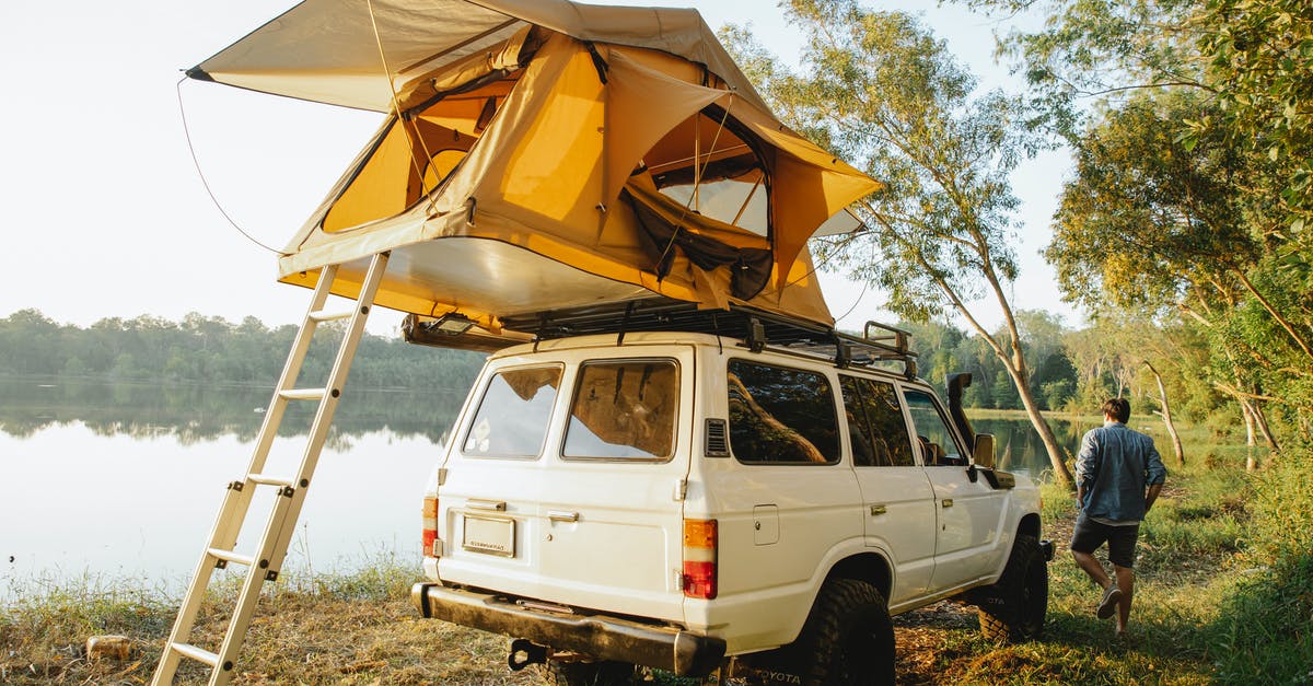 Is it legal to wild camp (with a car) in Ireland? - Back view of full body male traveler near yellow camping tent placed on roof of white car parked on coast of pond at daytime