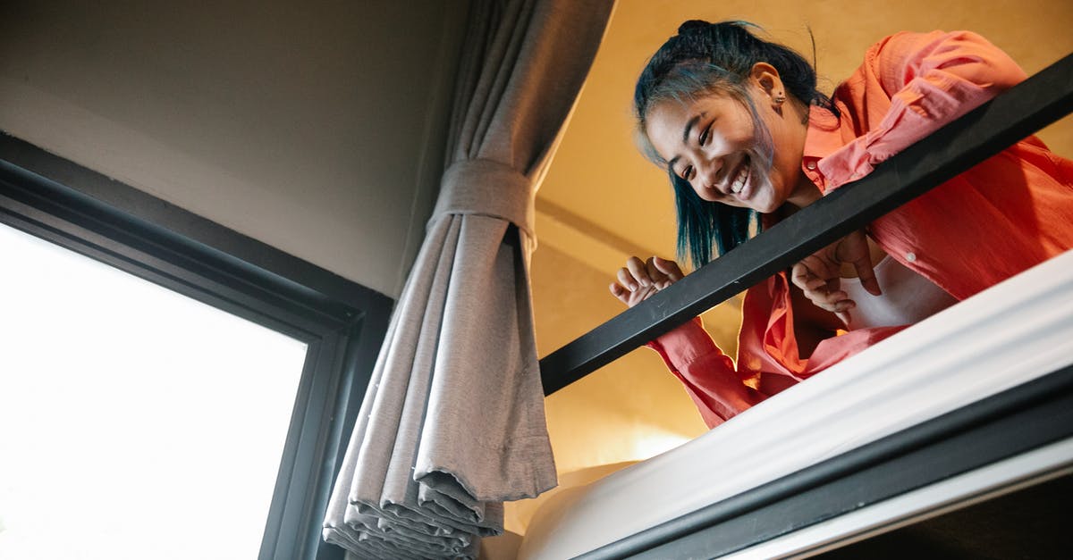 Is it possible to buy a travel curtain for hostels/trains? - Low angle of young ethnic cheerful female resting on bunk bed and looking down from top tier of bed near window with curtain